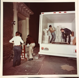 Two people loading cinder blocks onto a dolly while two other men are in the back of a large truck lifting cinder blocks to carry outside