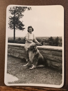 Photo of a woman and a collie standing outside. by a stone wall.
