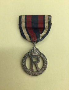 Photograph of a medal with white, red, and blue stripes on the ribbon and the letter R outlined in a circle as the medal