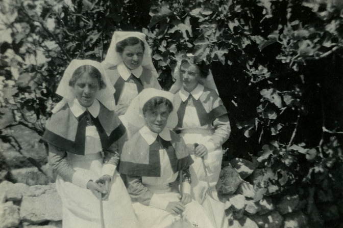 Black and white photograph of four women in nurses uniforms sitting and standing in front of a stand of fig trees