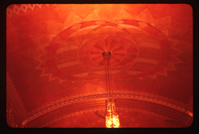 Photograph of orange and red tiles detailing on the ceiling above a small chandelier