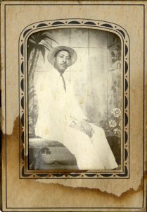 black and white mounted photograph of a man in a white dress suit sitting on a bench