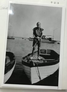 Black and white photograph of a woman standing in a row boat, holding an object, and smiling for the camera