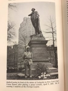 A man stands in uniform next to a statue of Lafayette with a bouquet of flowers on the ground next to him